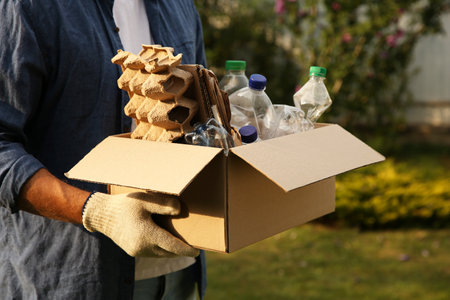 Recycling. Man holding cardboard box with different garbage outdoors, closeup