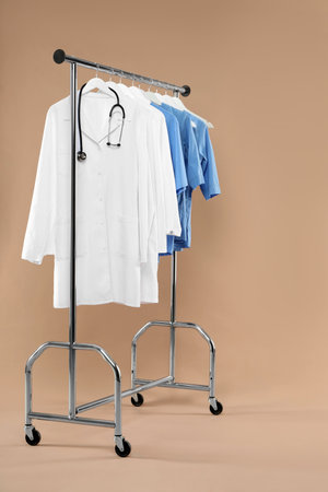 Different medical workers' uniforms and stethoscope on clothing rack against beige backgroundの写真素材