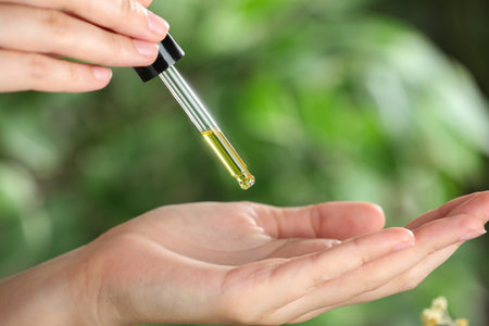 Woman dripping tincture from pipette onto hand against blurred green background, closeup