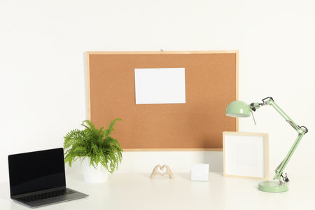 Cork board with blank paper note, laptop, lamp and decorative elements on light table near white wallの写真素材