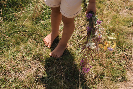 Little girl with bouquet of wildflowers walking barefoot outdoors on sunny day, closeup. Child enjoying beautiful natureの写真素材
