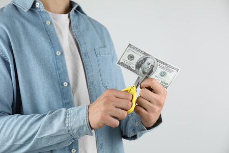 Budgeting. Man cutting dollar banknote with scissors on light gray background, closeupの写真素材