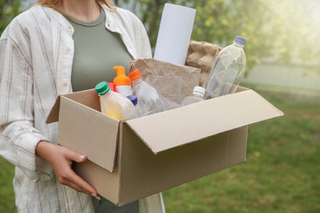 Recycling. Woman holding cardboard box with different garbage outdoors, closeupの写真素材