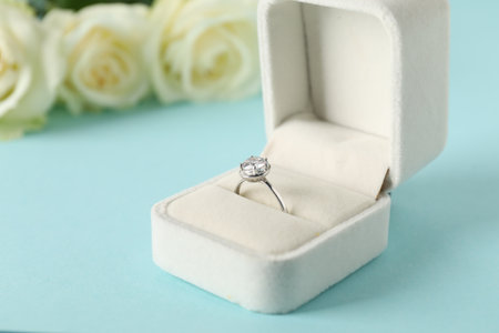 Beautiful ring with gemstone in box and flowers on light blue background, closeup