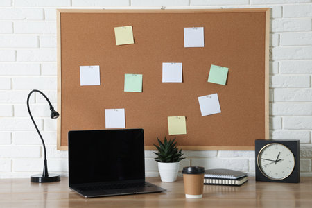 Cork board with blank paper notes, laptop, lamp, alarm clock and notebooks on wooden table near...