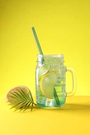Tasty refreshing drink with straw in mason jar, citrus fruit and palm leaf on yellow background