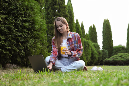 Woman with juice using laptop on green lawn in parkの写真素材