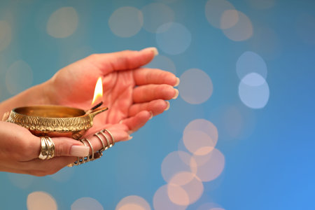 Diwali celebration. Woman holding lit diya lamp on light blue background with blurred lights, closeup. Space for textの写真素材
