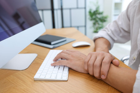 Man suffering from pain in wrist while working on computer at table indoors, closeup. Carpal tunnel...
