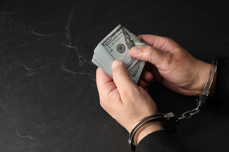 Corruption. Man in handcuffs holding dollar banknotes at gray textured table, closeup