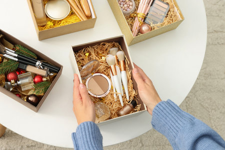 Woman holding Christmas gift boxes with makeup brushes and cosmetics at home, above view