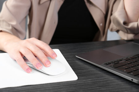 Woman using computer mouse while working with laptop at black wooden table, closeup