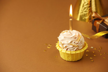 Birthday cupcake with burning candle, gift and party cone on brown background. Space for textの写真素材