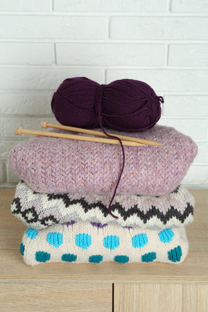 Skein of yarn, knitting needles and sweaters on wooden chest of drawers near white brick wall