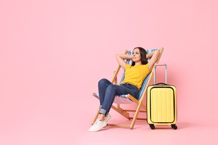 Happy young woman sitting on folding chair and suitcase against pink background, space for textの写真素材