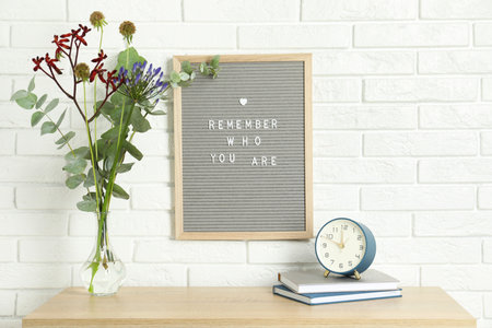 Letter board with phrase Remember Who You Are, books, alarm clock and bouquet in vase on wooden...