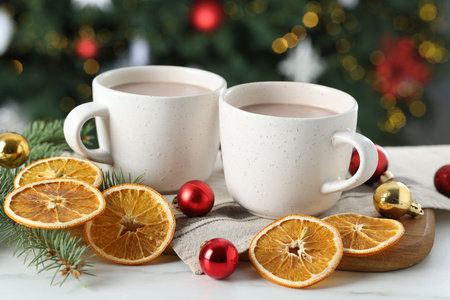 Tasty hot cocoa drinks in cups, fir branches, baubles and dry orange slices on white table against blurred lights, closeupの写真素材