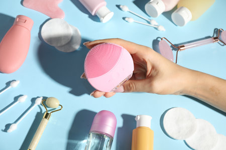 Woman holding facial cleansing brush near other body care products on light blue background, above...