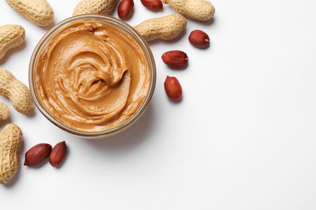 Tasty peanut butter in bowl and groundnuts on white table, flat lay. Space for text
