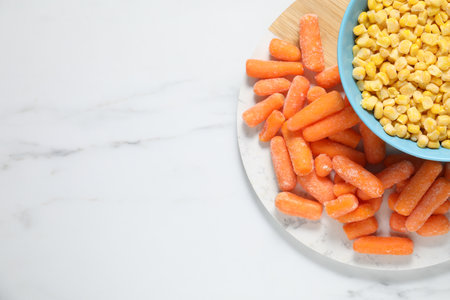 Frozen corn kernels in bowl and baby carrots on white marble table, top view. Space for textの写真素材