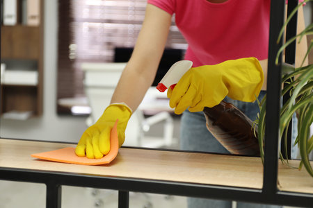 Young woman cleaning wooden shelf with rag and spray in office, closeup