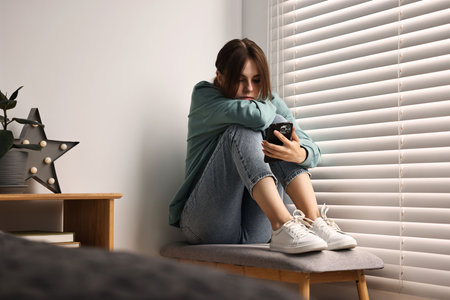 Loneliness concept. Sad teenage girl using smartphone at homeの写真素材