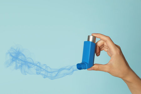 Asthma treatment. Woman holding inhaler on light blue background, closeup. Aerosolized medication coming out of deviceの写真素材