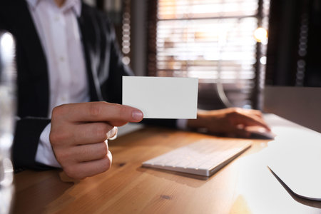Man holding blank business card while working with computer at table in office, closeup. Mockup for designの写真素材