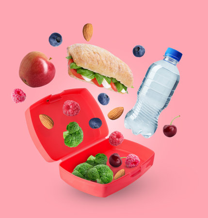 Different food and bottle of water falling into lunch box on pink backgroundの写真素材