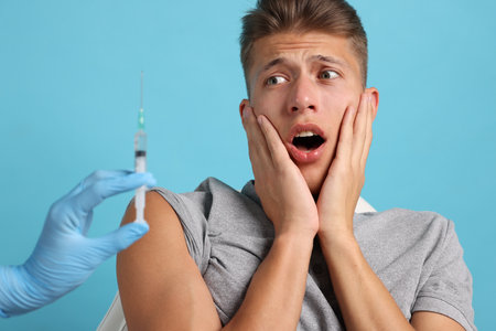Needle phobia. Doctor with syringe near scared man on light blue background, closeupの写真素材