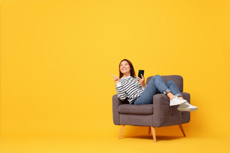 Smiling woman with smartphone sitting in armchair on yellow background. Space for textの写真素材
