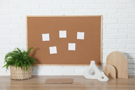Cork board with blank paper notes, laptop and decorative elements on wooden table near white brick wallの写真素材