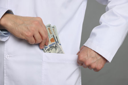 Corruption concept. Doctor putting dollar banknotes into his pocket on gray background, closeupの写真素材