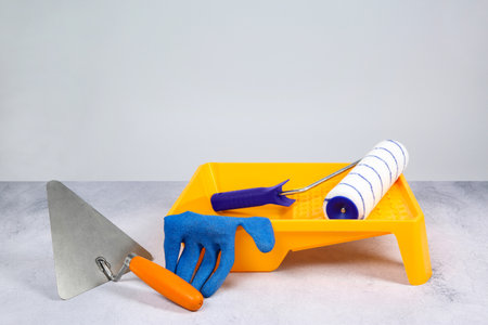 Paint roller, glove, trowel and yellow tray on textured table against light background. Building materialの写真素材