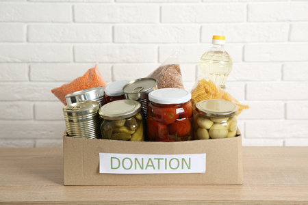 Different food products for donation in box on wooden table near white brick wallの写真素材