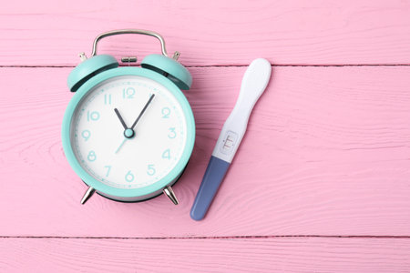 Pregnancy test and alarm clock on pink wooden table, top view. Space for text