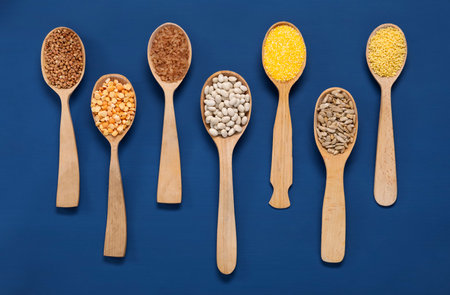 Different types of cereals, seeds and legumes in spoons on blue table, flat lay