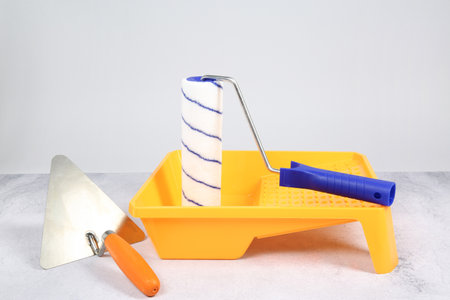 Paint roller, trowel and yellow tray on textured table against light background. Building material