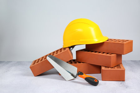 Red bricks, trowel and hardhat on textured table against light background