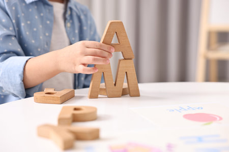 Little girl learning alphabet with wooden letters at white table indoors, closeup