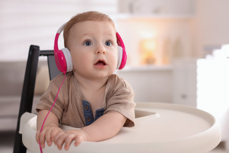 Cute little baby with headphones on high chair at home, space for textの写真素材