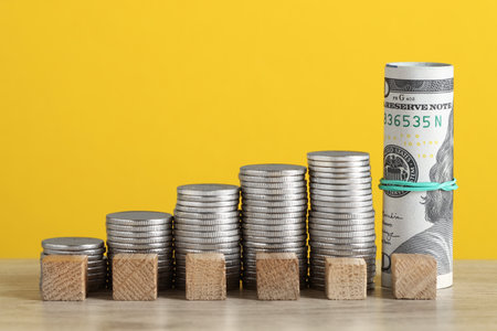 Blank cubes, stacked coins and dollar banknotes on wooden table against yellow background, closeup....