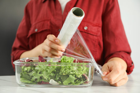 Woman putting plastic food wrap over glass container with salad at white table in kitchen, closeupの写真素材