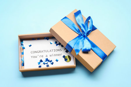 Card with phrase Congratulations You`re Winner in gift box on light blue background, above view