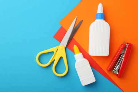 Glue, colorful paper, stapler and scissors on light blue background, flat lay. Space for text