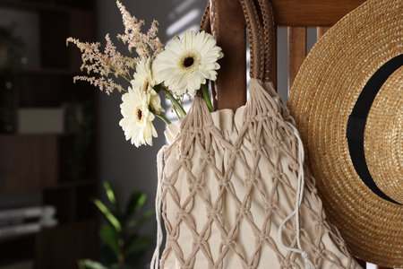 Handmade macrame shopping bag with flowers and straw hat on wooden chair indoorsの写真素材