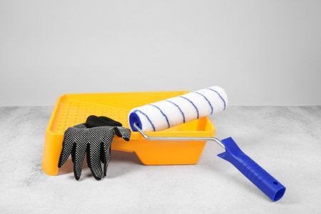 Paint roller, gloves and yellow tray on textured table against light background. Building materialの写真素材