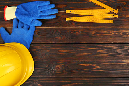 Yellow hard hat, rubber gloves and folding ruler on wooden table, flat lay. Space for text