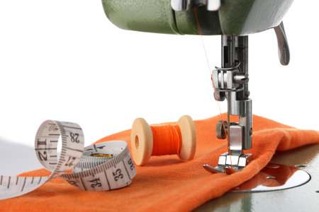 Sewing machine with orange fabric, measuring tape and spool of thread isolated on white