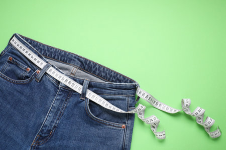 Jeans and measuring tape on light green background, top view. Space for text